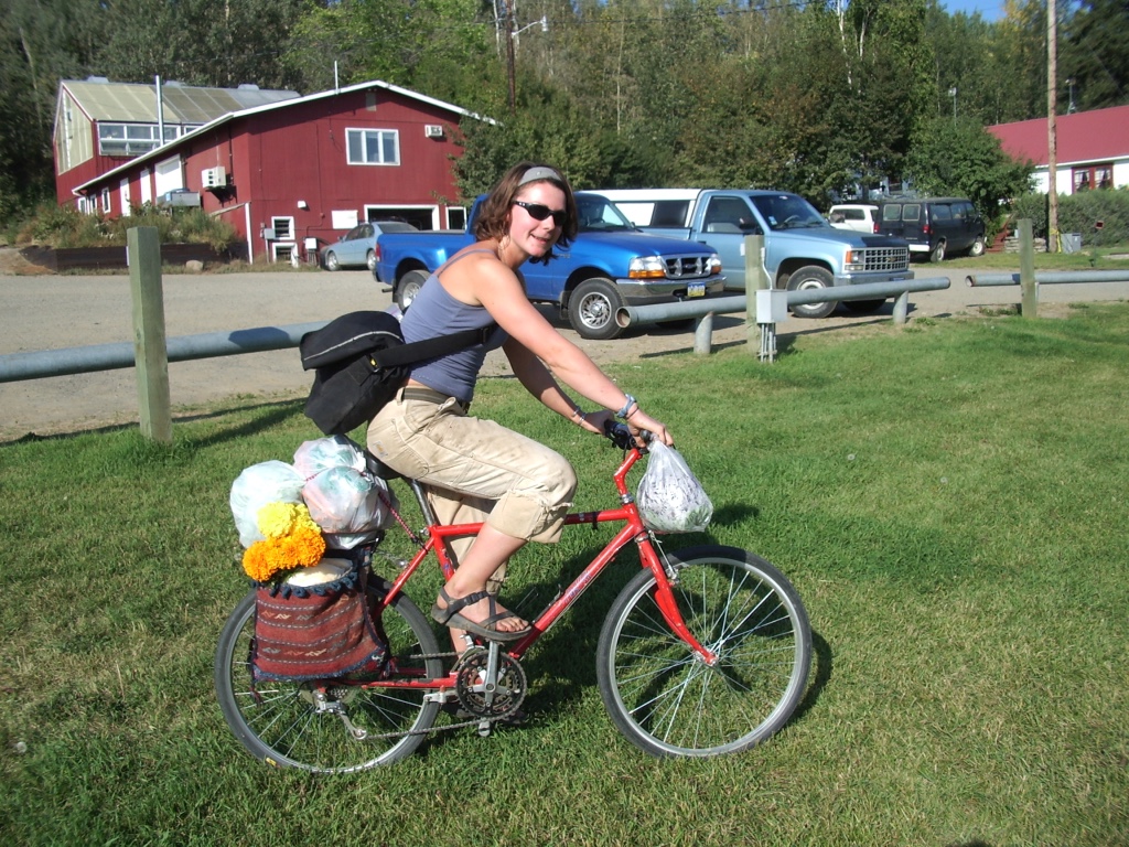heidi on red bike with flowers and vegetables strapped on at farm with cars in the background